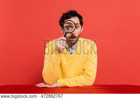 Scrupulous Man In Yellow Long Sleeve Jumper Holding Magnifying Glass On Red Coral Background.
