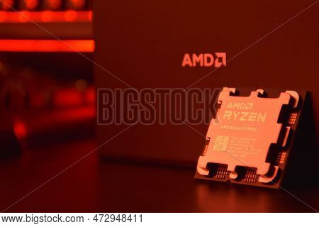 A Close-up Of An Amd Ryzen 9 7900X Processor In A Packaging Box With Backlit Keyboards In The Backgr