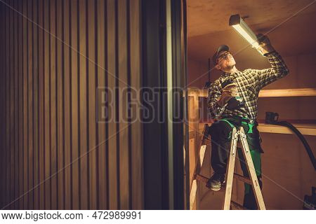 Caucasian Electrician In His 40s Installing Led Light Fixture In The Shed Ceiling. Small Wooden Buil