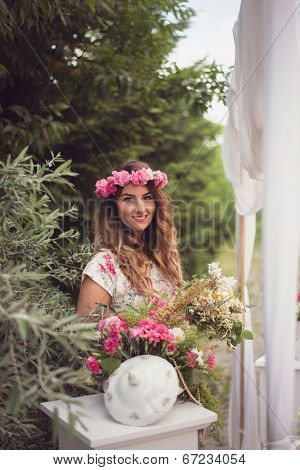 Beautiful Woman With A Crown Of Flowers On Her Head, Sitting Behind A Flowerpot And Holding A Bouque