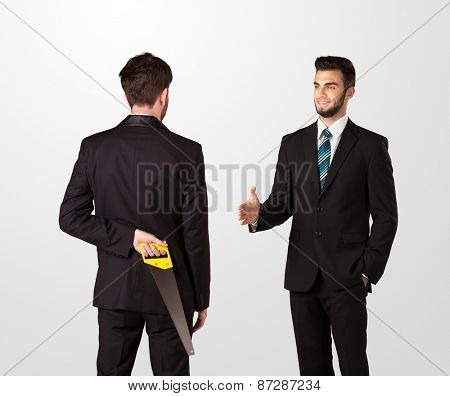 Two opposing businessman shake hands, one of them hiding a weapon behind his back