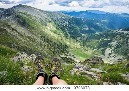Somebody is sitting at the edge of cliff in mountains, showing his feet wearing hiking shoes. First person shoot. Retezat area, Carpathians, Romania