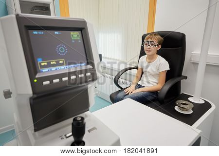 Boy in ophthalmic glasses sits in armchair in medical room.