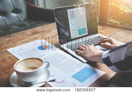 Closeup business woman hands using laptop with cup of coffee graph financial and network diagram documents on wooden table.