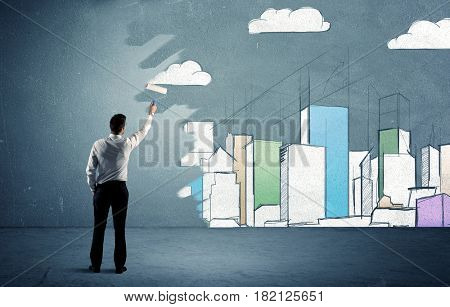 An elegant businessman standing with a paint roller transforming a blue wall into urban city landscape drawing sketch including skyscrapers and clouds concept