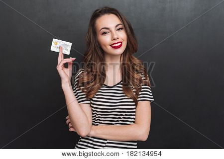 Image of smiling young lady standing over grey wall and holding debit card in hands. Looking at camera.