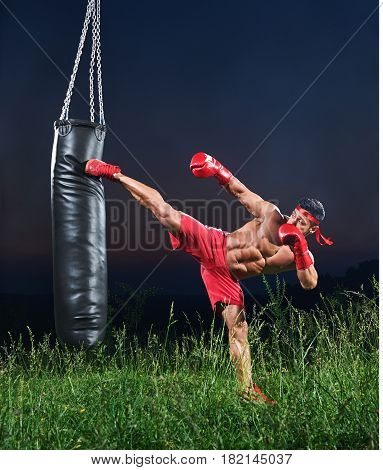Handsome young strong muscular fighter exercising with a punching bag practicing his high kick martial arts combat fighting kickboxing Muay Thai strength power masculinity energy effort athlete.