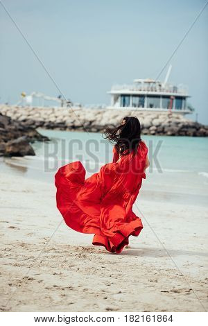 Beautiful young woman with long hair in red dress goes along the beach. Girl in red dress. Long red dress flutters in the wind