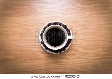 White cup with black, hot coffee standing on brown, roasted beans on wood table, close up.