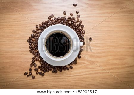 White cup with black, hot coffee standing on brown, roasted beans on wooden table, close up