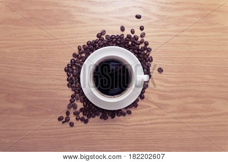 White cup with black, hot coffee standing on brown, roasted beans on wood table, close up