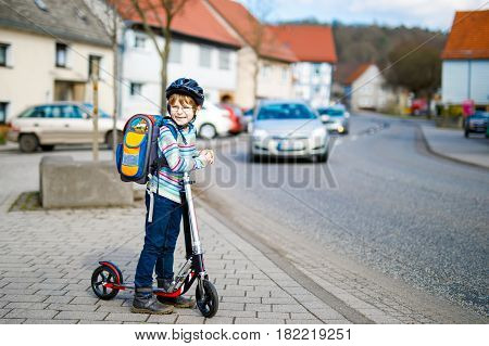 Active school kid boy in safety helmet riding with his scooter in the city with backpack on sunny day. Happy child in colorful clothes biking on way to school. Safe way for kids outdoors to school.