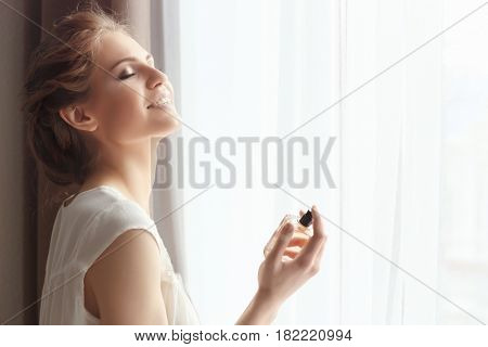 Beautiful young woman with bottle of perfume near window at home