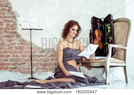 Beautiful woman sitting on the floor near the chair with violin in case and holding notes in hand
