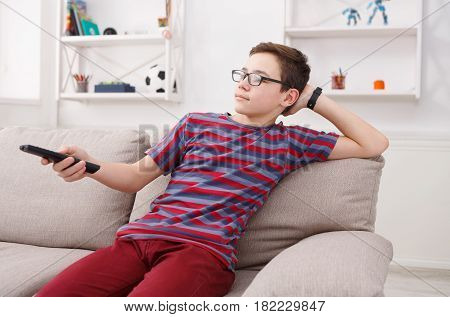 Teenager boy watching television, using TV remote while sitting on couch in living room at home