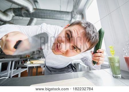 Young male chef in uniform cutting cucumber and looking crazily at camera while having fun.