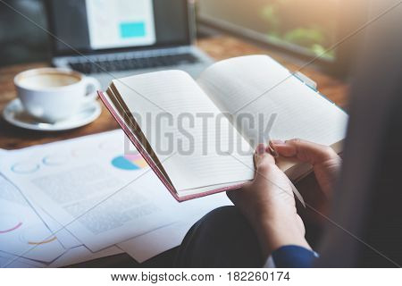 Closeup business woman hands holding notebook. Graph financial diagram documents laptop and cup of coffee on wooden table.