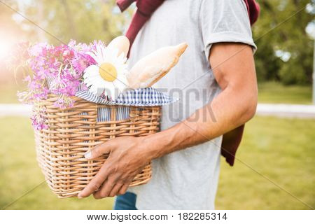 Closeup of basket with food, drinks and flowers for picnic holded by young man in park