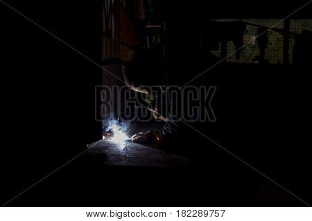 Worksman welding wrought iron metal rods together in dark workshop with blue smoke and orange sparks wearing protective UV eyewear gear for eye protection