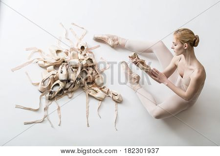 Young ballerina in white tights is sitting on the floor and holding a ballet slip in her hands. Before her a pile of pointe shoes in different condition. Studio shooting, horizontal top shot.