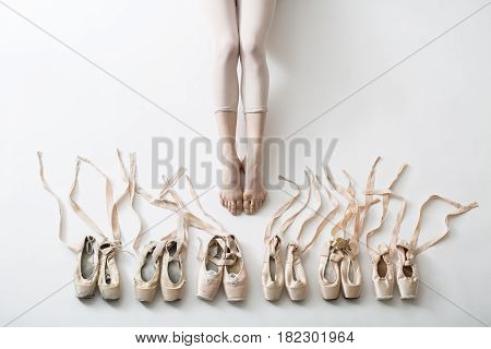Many pairs of ballet shoes in pairs stand in a row. Legs of a young ballerina with bare feet. The girl's fingers are all covered in wounds and plasters. Pointe shoes in different condition from new to very shabby old. Tapes are accurately laid out in diff