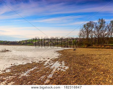 The flat river bank Kuban covered with gravel and ahead on a of the house and the blossoming trees. The blue sky with white clouds.
