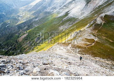 Hiker with backpack in distance on picturesque highland landscape standing on curvy trail over steep mountain shot from above. Pirin mountain under the peak Vihren Bulgaria