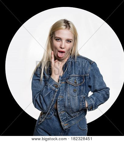 young woman in denim jaket and jeans shows tongue between two fingers on white background. indecent lifestyle concept. White circle on a black background