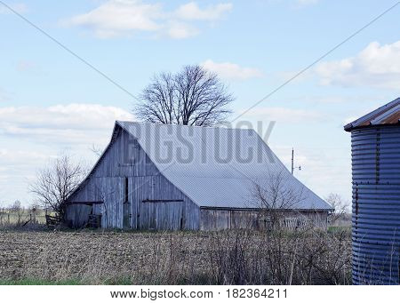 Old barn with single grain bin in foreground, open field in the foreground, blue skies with white clouds