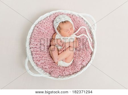 Adorable kid napping on big pink pillow wearing a hat and cute suit, in basket