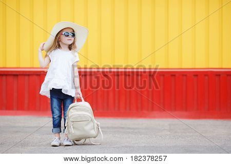 Beautiful little girl 6-8 years old,in a large straw hat from the sun,dressed in a short white dress without sleeves and blue jeans,with long blonde wavy hair and wears dark sun glasses in blue frame,posing standing near the school with a white backpack