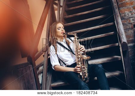 Playing young attractive girl sitting on steps in white shirt with a saxophone - outdoor in old town. Sexy young woman with sax.