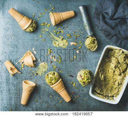 Homemade pistachio ice cream in ceramic mold and metal scooper with crashed pistachio nuts and waffle cones over grey concrete background, top view, copy space