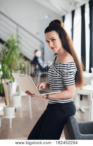 Beautiful adult female is holding a laptop and looking at the camera while standing in an empty cafe