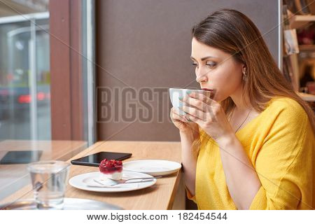 A young woman is taking a sip of coffee at the table with raspberry cake, glass of water and tablett on it. The scene is next to a window.