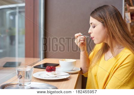 A young woman eating delicious raspberry cake at the table next to a window with cup of coffee, glass of water and tablett on it.