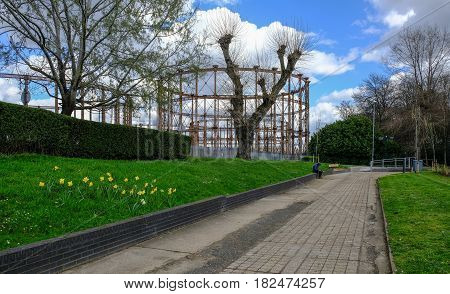 Disused gasworks in East London from pathway near River Lea. Shows a man sitting on the wall. A springtime shot with clouds in the sky.