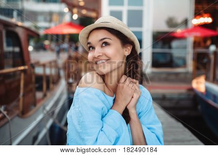 Portrait of beautiful smiling white Caucasian brunette girl on boat yacht pier quay in blue dress and straw hat posing making funny emotional faces expressions having fun