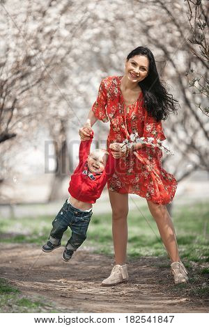 Young mother with her baby on walk in blooming garden. They play and laugh cheerfully among flowering apricot trees.