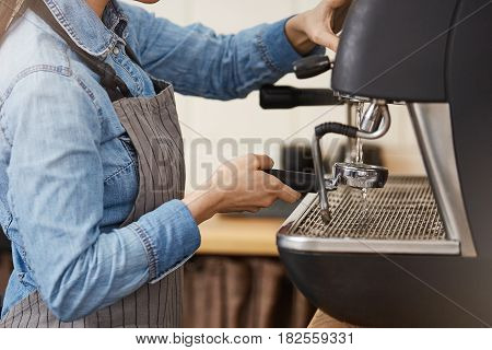 Closeup of female bartender in denim shirt and apron cleaning bottomless portafilter with steamer.