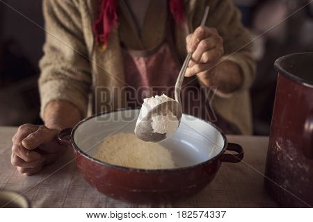 Detail of an elderly woman's hand holding a ladle and adding some flour into a bowl in preparation for making homemade pasta. Selective focus