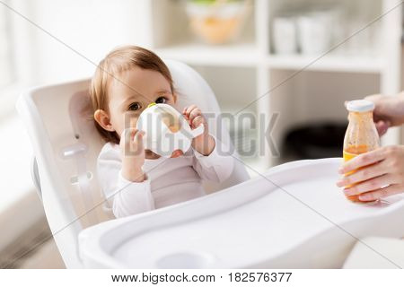 childhood and people concept - little baby drinking from spout cup sitting in highchair at home