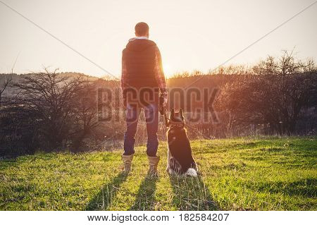 A man with a beard walking his dog in the nature, standing with a backlight at the rising sun, casting a warm glow and long shadows against the background of the gorge and trees on caucazus