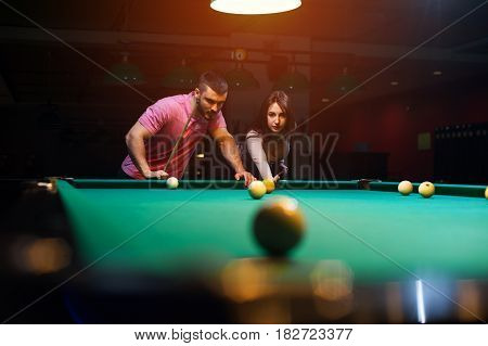 Young romantic couple having fun playing billiard game in dark club. Young brunette girl aiming to take a snooker shot, while man standing next to her and looking at table.