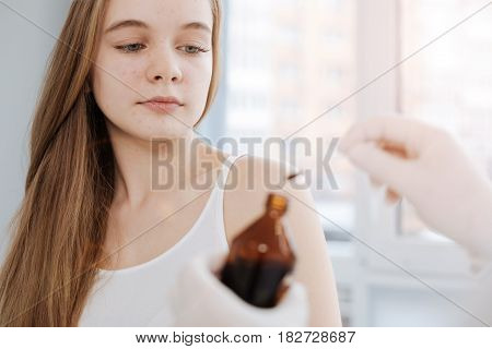 Useful nature of iodine. Fearless motionless charming teenager sitting in the hospital and waiting for treatment while dermatologist holding the bottle of iodine and cotton swab