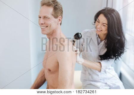 Checkup of cancerous moles. Joyful cheerful experienced specialist working in the clinic and enjoying appointment while examining man back and using dermatoscope