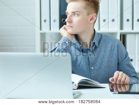 Handsome pensive young business man sitting at the dask with laptop and looking at the window.