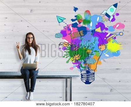 Young woman with laptop sitting on table and showing peace sign on woodwn background with colorful sketch. Creative start up concept