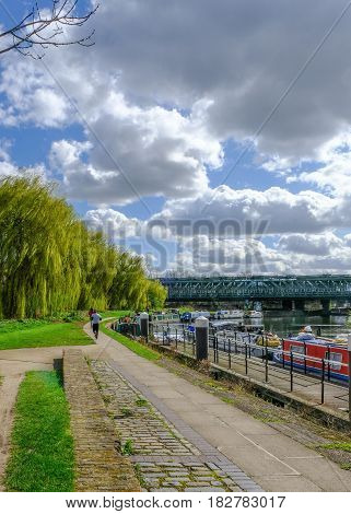 Towpath with joggers by River Lea Bow London. Shows willow trees and railway bridge in spring.