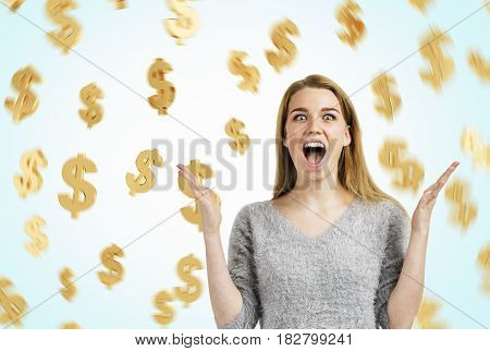 Portrait of a happy blond woman wearing a gray sweater screaming with joy while standing against a blue wall with dollar signs